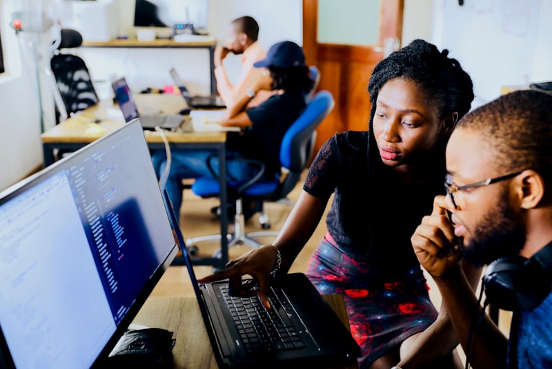 Two colleagues talking across a desk in a bright office, one gesturing while explaining.